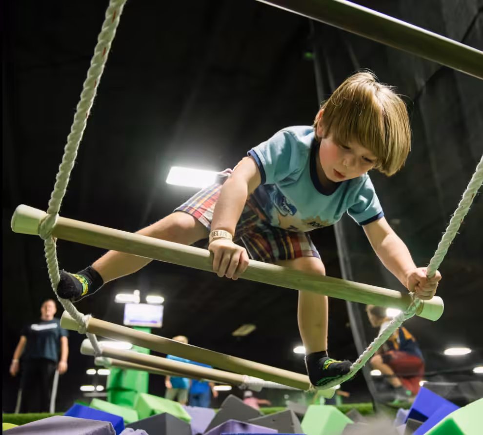 A young boy climbs across a suspended wooden rope ladder over a foam pit at an indoor Trampoline Park, focusing intently as he balances with his arms and legs on the rungs. Other people are visible in the background.