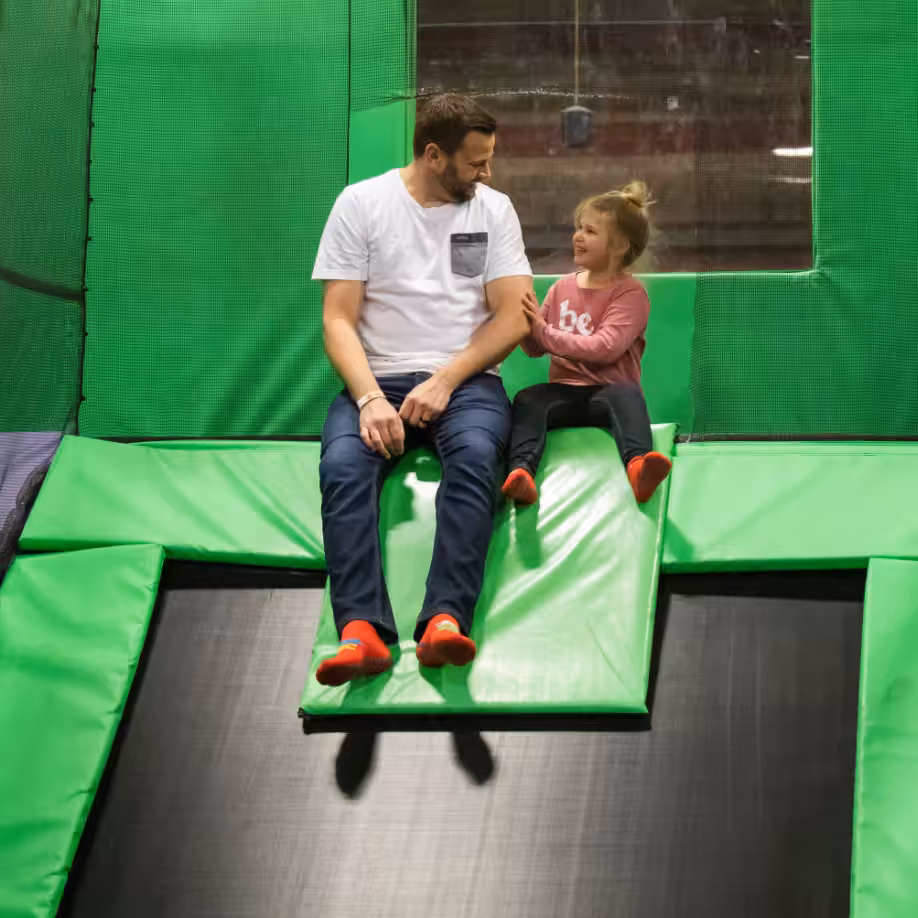 A man and a young girl wearing red socks sit on a green and black trampoline slide, smiling and talking to each other in an indoor play center.