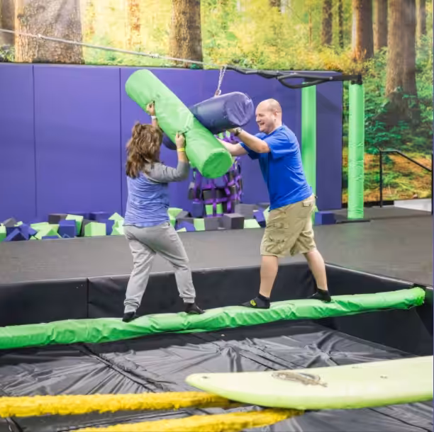 Two people balance on a padded beam indoors, playfully jousting with large foam batons. The background features a forest wall mural and a pit filled with foam blocks.
