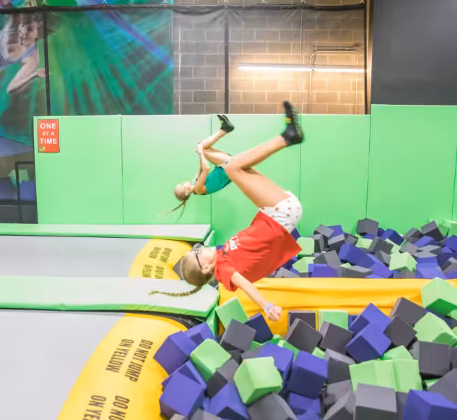 Two children are doing backflips into a foam pit at an indoor trampoline park. The pit is filled with blue, gray, and green foam cubes, and the area is surrounded by green padding and safety signs.