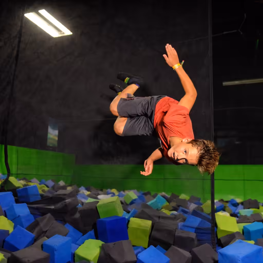 A boy wearing a red shirt and gray shorts is mid-air doing a flip above a foam pit filled with blue, green, and black foam blocks at a trampoline park.