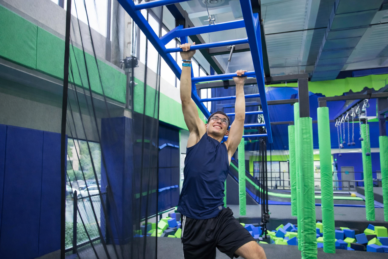 A man in athletic wear hangs from blue monkey bars at an indoor trampoline park, smiling as he moves between bars. The background features foam pits and green padded pillars.