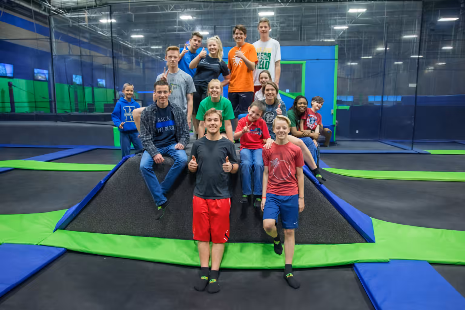 A group of teens smiles and poses together on a padded structure at a lively trampoline park, surrounded by blue and green mats.