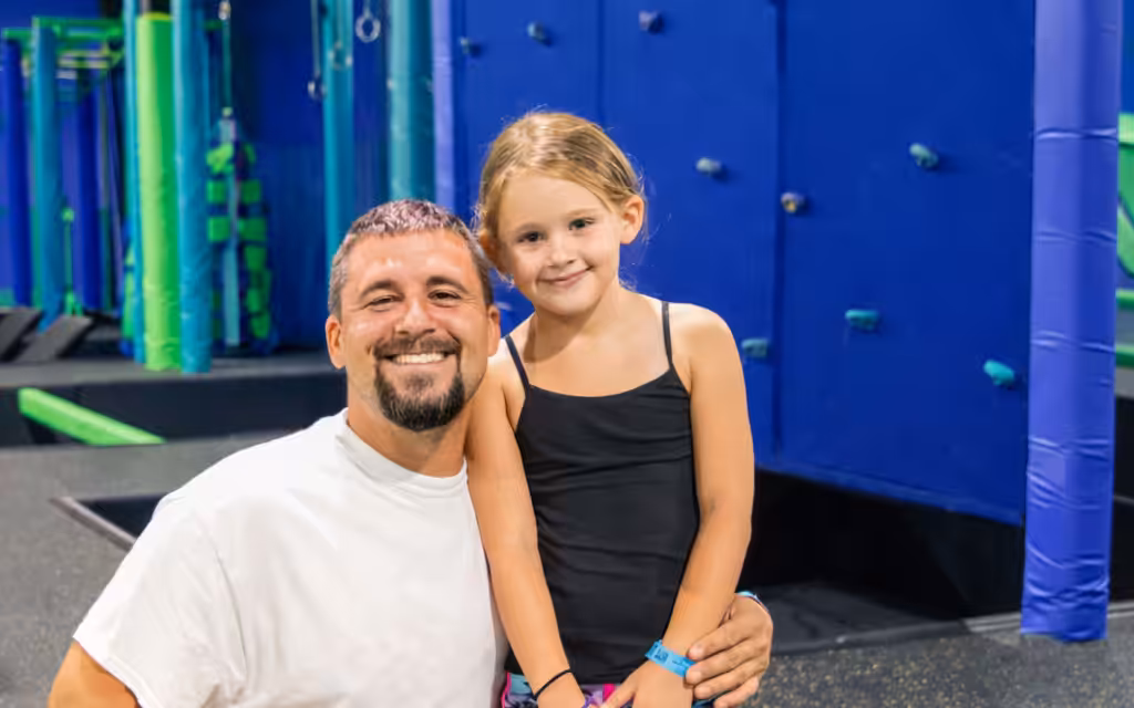 A smiling man in a white t-shirt kneels next to a young girl in a black tank top at an indoor trampoline park with blue and green padded walls and equipment in the background.