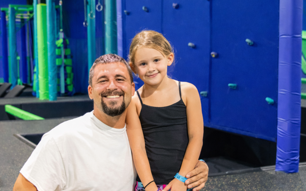 A smiling man in a white t-shirt kneels next to a young girl in a black tank top at an indoor trampoline park with blue and green padded walls and equipment in the background.