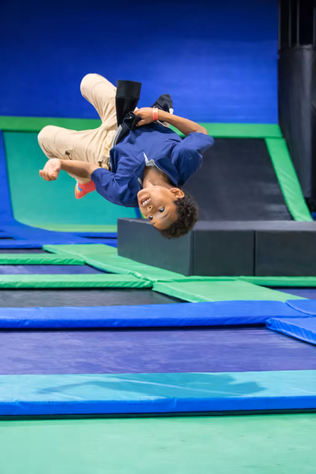 A young boy mid-backflip at a vibrant trampoline park, with blue and green padded mats and walls in the background. He looks excited and focused as he spins through the air.