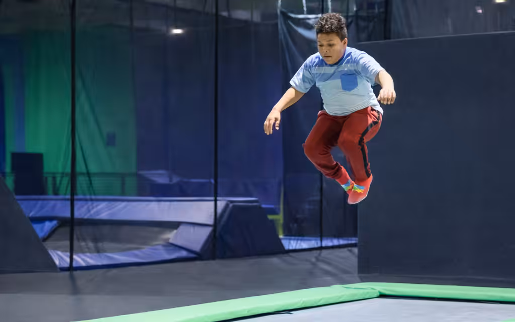 A boy wearing a light blue shirt and red pants is midair, jumping on a trampoline at an indoor trampoline park with padded walls and safety nets.