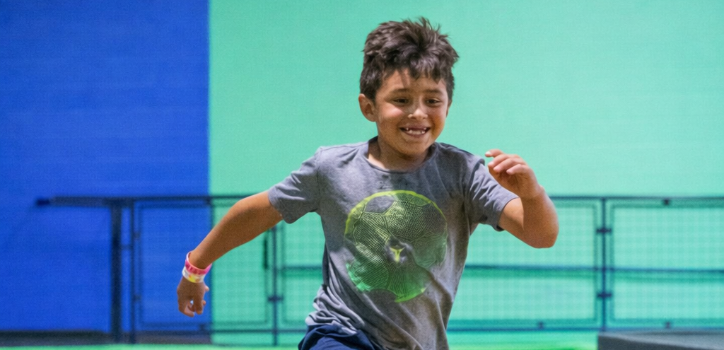 A young boy wearing a gray t-shirt with a green soccer ball design runs indoors, smiling, against a blue and green wall background at a trampoline park.