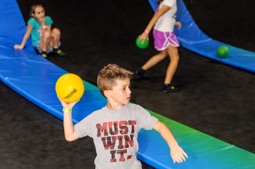 A boy in a MUST WIN IT. shirt prepares to throw a yellow ball at an indoor trampoline park, while two girls and several green balls are visible in the background on blue padded ramps.