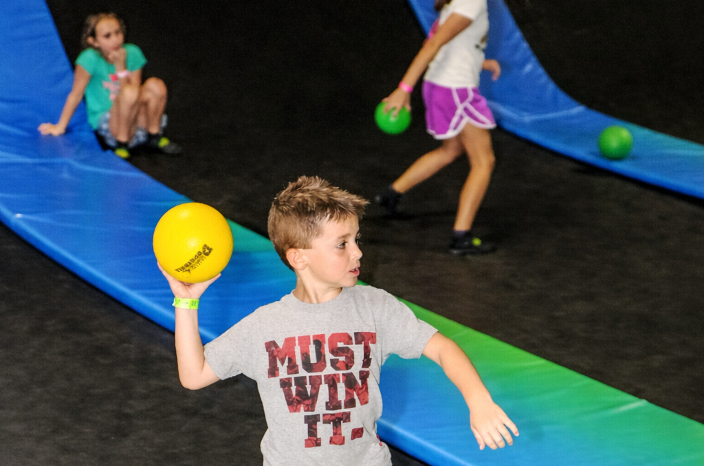 A boy in a MUST WIN IT. shirt prepares to throw a yellow ball at an indoor trampoline park, while two girls and several green balls are visible in the background on blue padded ramps.