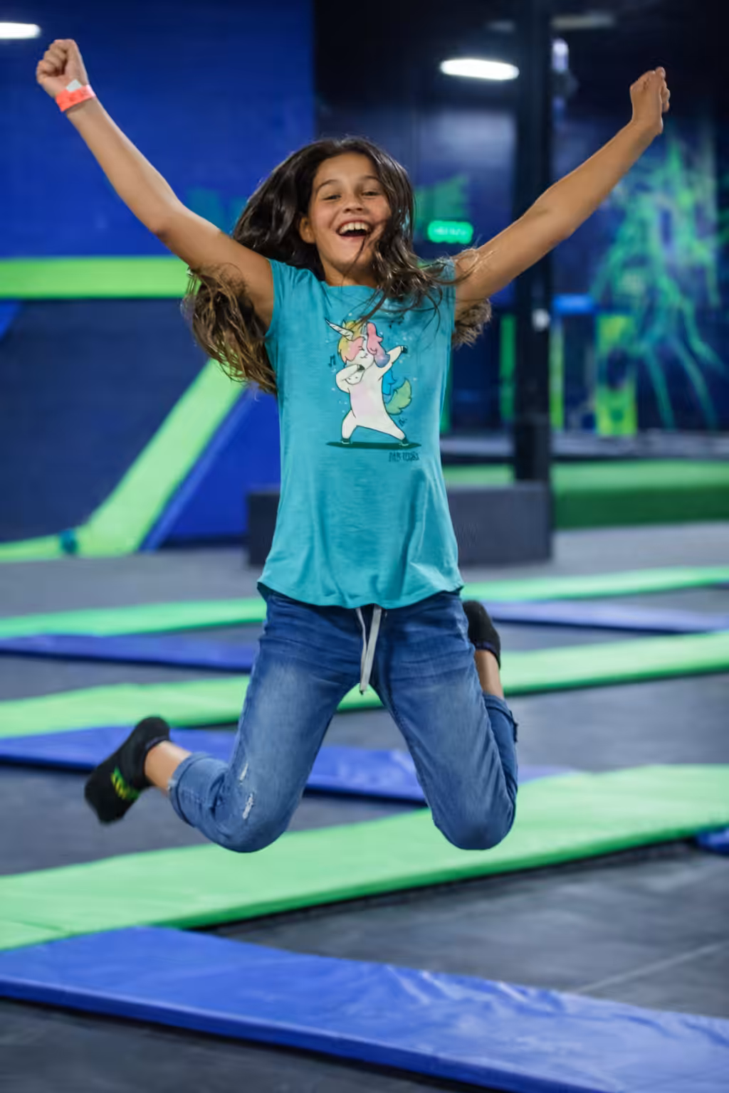 A smiling girl with long hair jumps joyfully at a trampoline park, wearing a blue shirt with a cartoon unicorn, blue jeans, and black socks. The brightly colored trampoline areas are blue and green.
