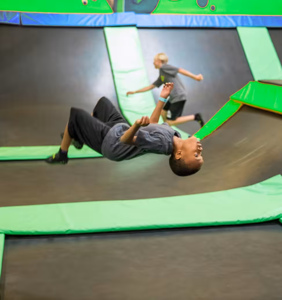 A boy in a gray shirt does a backflip on a trampoline at a bustling trampoline park, while another child runs in the background. Bright green padding outlines the trampolines.