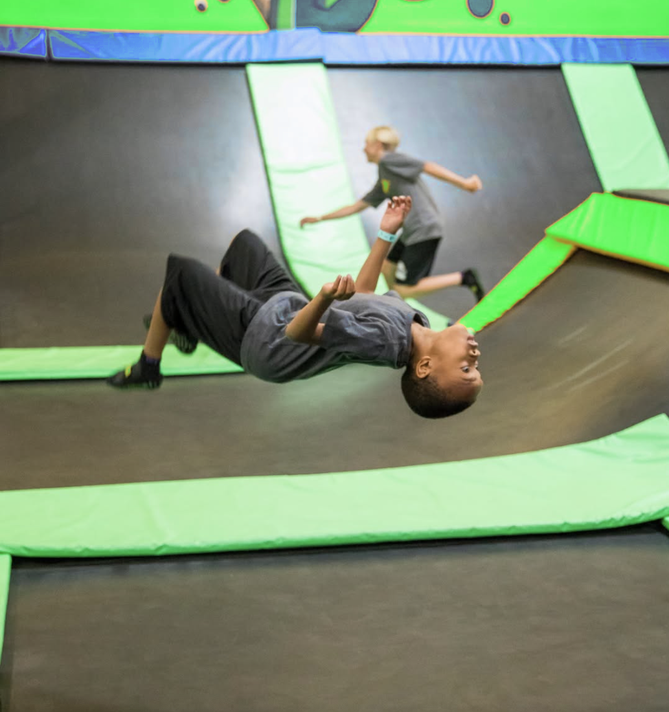 A boy in a gray shirt does a backflip on a trampoline at a bustling trampoline park, while another child runs in the background. Bright green padding outlines the trampolines.