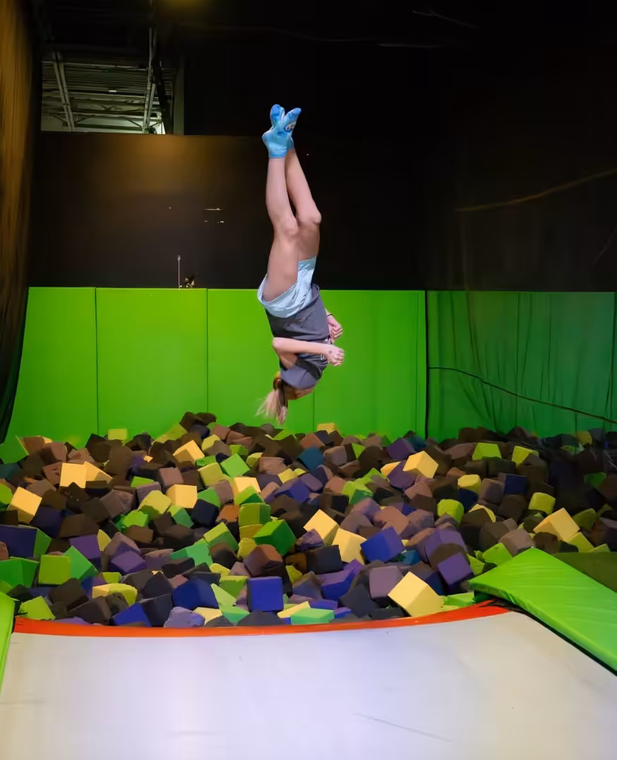 A person is upside down in mid-air, performing a flip above a foam pit filled with colored blocks at a lively trampoline park.