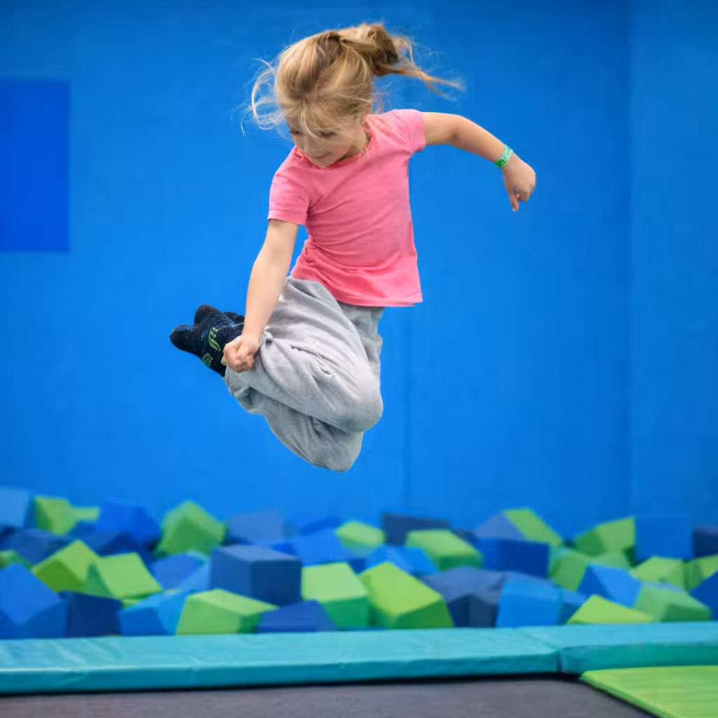 A young girl in a pink shirt and gray sweatpants jumps mid-air above a trampoline at a lively trampoline park, with foam blocks in blue and green colors in the background.